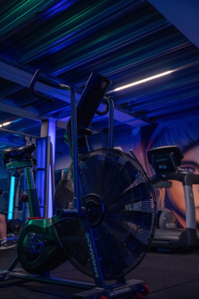 A modern gym filled with exercise equipment under blue lighting, featuring a stationary fan bike.